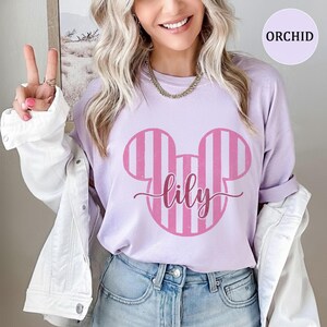 K&ouml;nnte beinhalten: Ein hellviolettes T-Shirt mit einer gro&szlig;en, rosa gestreiften Micky-Maus-Ohren-Silhouette und dem Namen lily in Schreibschrift in der Mitte. Das T-Shirt wird unter einer wei&szlig;en Jeansjacke mit silbernen Kn&ouml;pfen und sichtbaren N&auml;hten getragen.