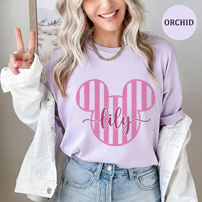 K&ouml;nnte beinhalten: Ein hellviolettes T-Shirt mit einer gro&szlig;en, rosa gestreiften Micky-Maus-Ohren-Silhouette und dem Namen lily in Schreibschrift in der Mitte. Das T-Shirt wird unter einer wei&szlig;en Jeansjacke mit silbernen Kn&ouml;pfen und sichtbaren N&auml;hten getragen.