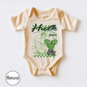 Pu&ograve; includere: Un body per neonati a maniche corte di colore naturale con una stampa grafica di Hulk, il testo "Hulk" e "2008" sopra un motivo a scacchi verde e bianco. Il design include una grande illustrazione verde del personaggio sul davanti.