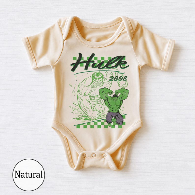 Pu&ograve; includere: Un body per neonati a maniche corte di colore naturale con una stampa grafica di Hulk, il testo "Hulk" e "2008" sopra un motivo a scacchi verde e bianco. Il design include una grande illustrazione verde del personaggio sul davanti.
