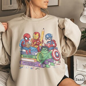 Puede incluir: Una sudadera de cuello redondo color arena con un estampado gr&aacute;fico de superh&eacute;roes de dibujos animados, incluyendo a Spider-Man, Iron Man, Captain America y Hulk, sentados sobre una pila de libros. Los libros est&aacute;n etiquetados como Super Physics, World History y Avengers Assemble.