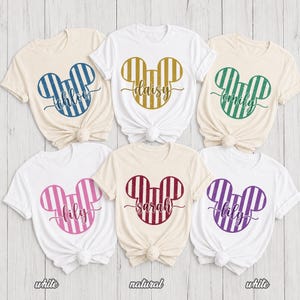 K&ouml;nnte beinhalten: Sechs kurz&auml;rmelige T-Shirts in Wei&szlig; und Naturfarben, jedes mit einer gro&szlig;en Mausohr-Silhouette mit vertikalen Streifen in Blau, Gold, Gr&uuml;n, Pink, Kastanienbraun oder Lila, wobei ein Name in Schreibschrift &uuml;ber die Mitte gedruckt ist.