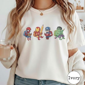 May include: An ivory t-shirt featuring a graphic print of four cartoon superheroes. Spider-Man reads a book, Iron Man holds a large pencil, Captain America reads a book, and the Hulk holds a large pencil. Each character wears a backpack.