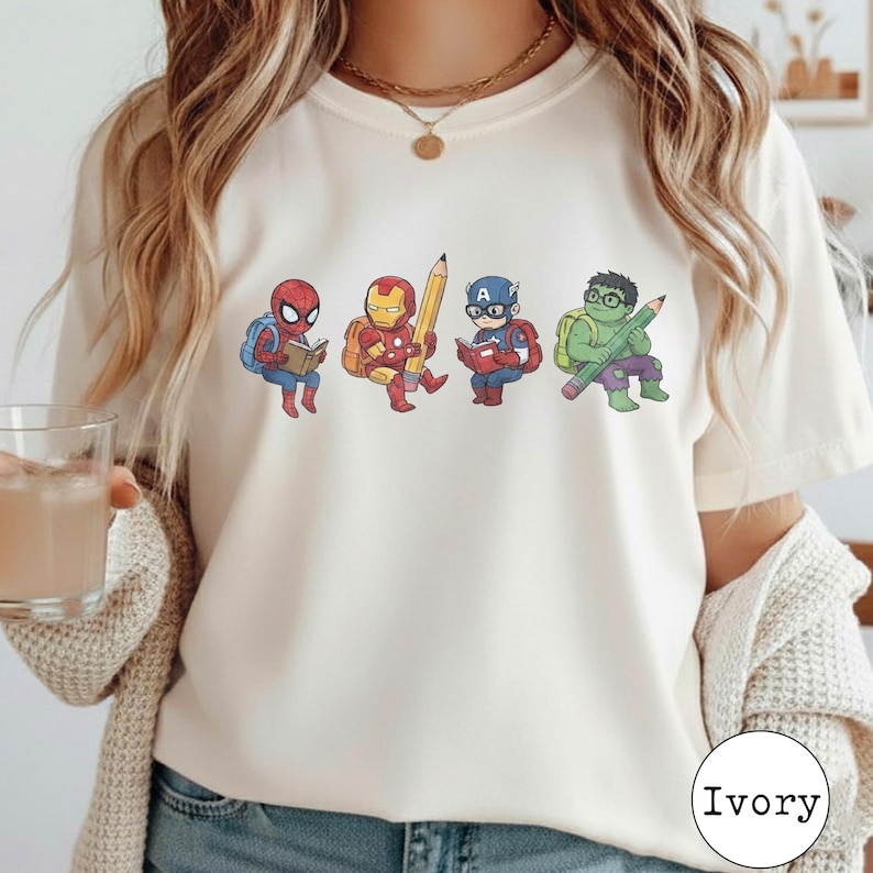 May include: An ivory t-shirt featuring a graphic print of four cartoon superheroes. Spider-Man reads a book, Iron Man holds a large pencil, Captain America reads a book, and the Hulk holds a large pencil. Each character wears a backpack.