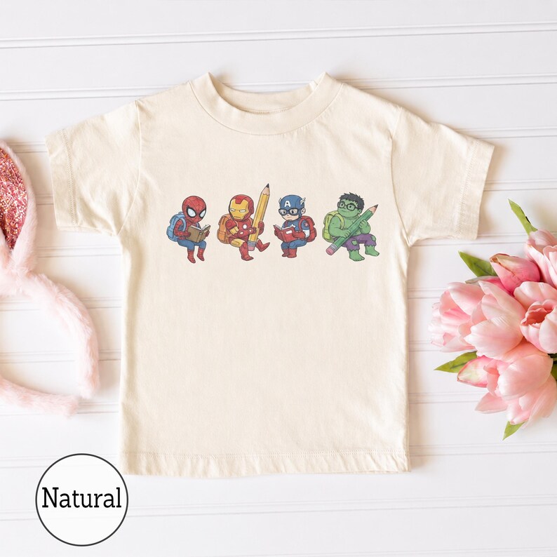 May include: A natural-colored short-sleeve t-shirt featuring a graphic print of four cartoon superheroes. The characters include Spider-Man reading a book, Iron Man holding a large pencil, Captain America reading, and the Hulk holding a pencil.