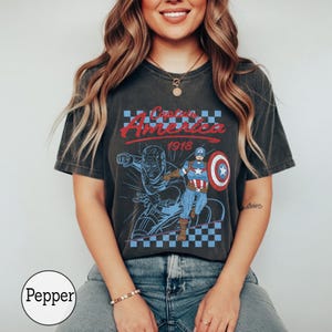 Pu&ograve; includere: Una t-shirt grigio antracite a maniche corte con una grafica di Captain America e la scritta Captain America 1918. Il design include un motivo a scacchi blu e bianco, uno scudo rosso e un'illustrazione in stile vintage del personaggio.