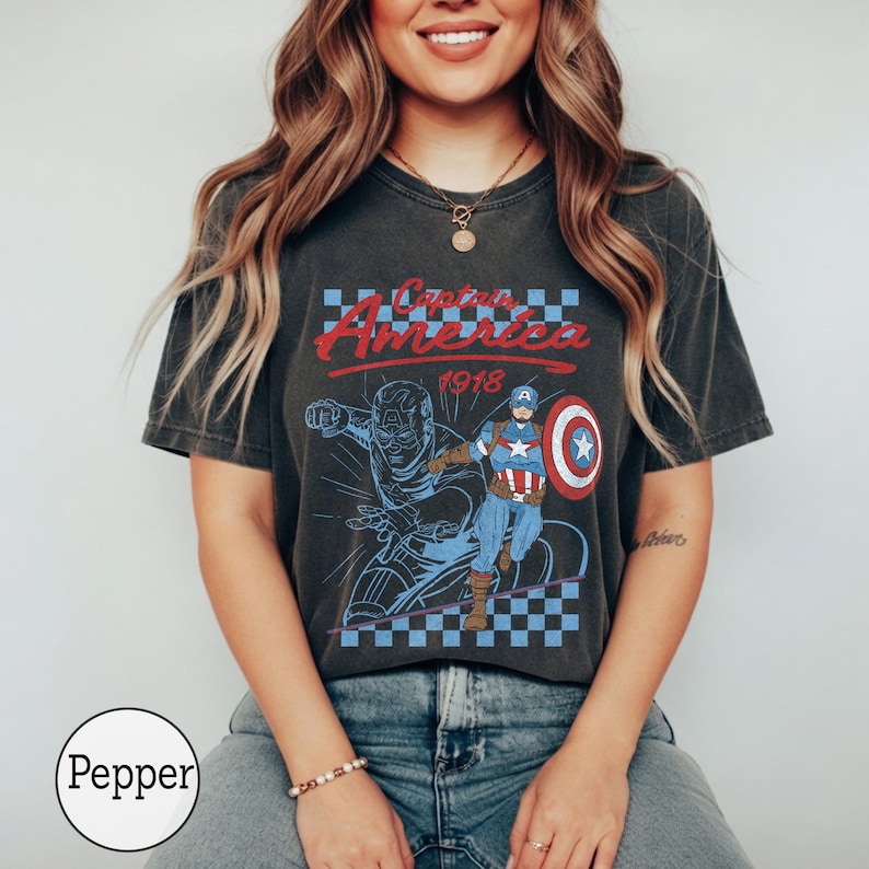Pu&ograve; includere: Una t-shirt grigio antracite a maniche corte con una grafica di Captain America e la scritta Captain America 1918. Il design include un motivo a scacchi blu e bianco, uno scudo rosso e un'illustrazione in stile vintage del personaggio.