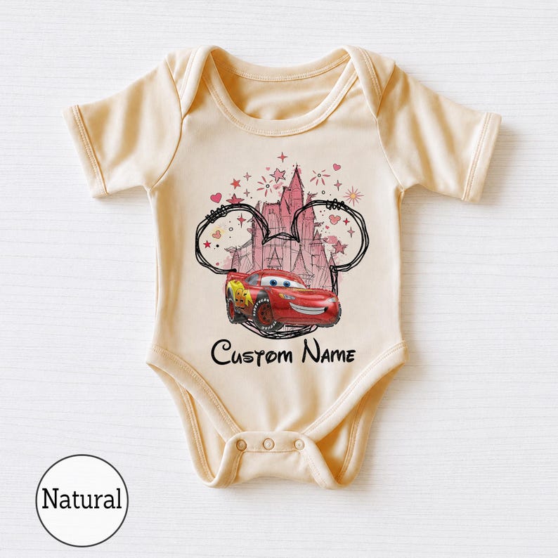 K&ouml;nnte beinhalten: Ein cremefarbener Kurzarm-Babybody mit einer Grafik eines roten Rennwagens vor einer rosa Schloss-Silhouette, umrahmt von einer schwarzen Umrisszeichnung von Mausohren, Sternen und Herzen, mit dem darunter gedruckten Text Custom Name in Schwarz.