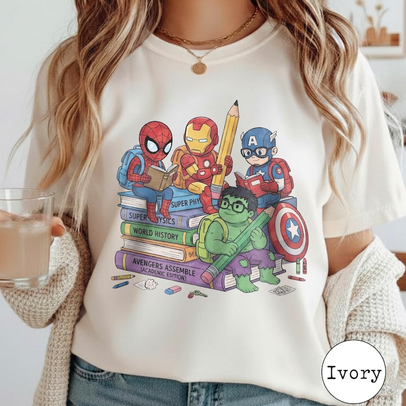 Puede incluir: Una camiseta de color marfil con un estampado gr&aacute;fico de superh&eacute;roes de dibujos animados, incluidos Spider-Man, Iron Man, Capit&aacute;n Am&eacute;rica y Hulk, sentados sobre una pila de libros titulados Super Physics, World History y Avengers Assemble Academic Edition.