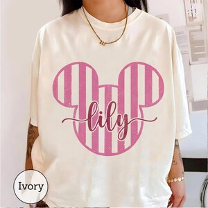 K&ouml;nnte beinhalten: Ein elfenbeinfarbenes Kurzarm-T-Shirt mit einer gro&szlig;en Grafik einer Mausohren-Silhouette in Rosa und Wei&szlig; auf der Brust. Der Name lily ist in einer dunkelrosa Schreibschrift in der Mitte des Mausohren-Designs aufgedruckt.