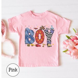 May include: A light pink short-sleeve t-shirt featuring a graphic print of the word BOY in large letters. The letters are decorated with patterns and images of cartoon race cars, a checkered flag, and a Route 66 sign. Small stars are scattered.