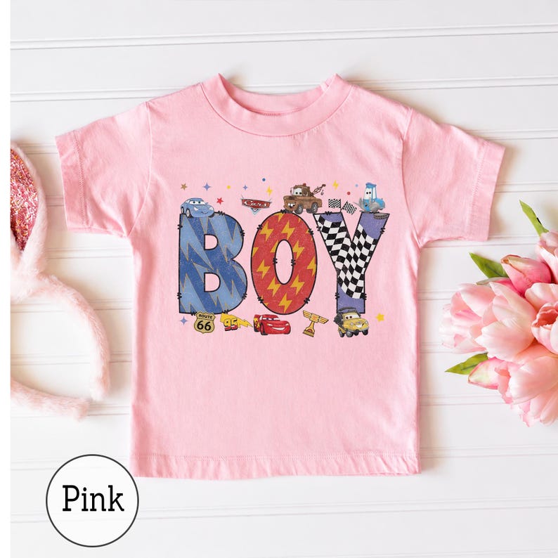 May include: A light pink short-sleeve t-shirt featuring a graphic print of the word BOY in large letters. The letters are decorated with patterns and images of cartoon race cars, a checkered flag, and a Route 66 sign. Small stars are scattered.