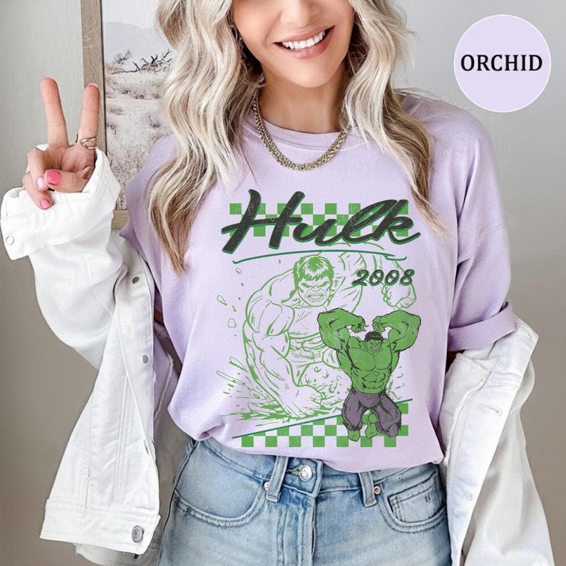 Pu&ograve; includere: Una t-shirt color viola chiaro con una grafica di Hulk e il testo Hulk e 2008. Il design include un motivo a scacchi verde e un disegno al tratto del personaggio. La t-shirt &egrave; indossata sotto una giacca di jeans bianca con bottoni argentati.