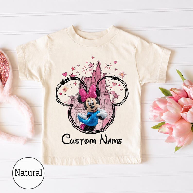 May include: A natural-colored short-sleeve t-shirt featuring a graphic of Minnie Mouse in a blue dress and pink bow, centered within a black hand-drawn style outline of mouse ears. Behind the character is a pink castle and the text Custom Name.