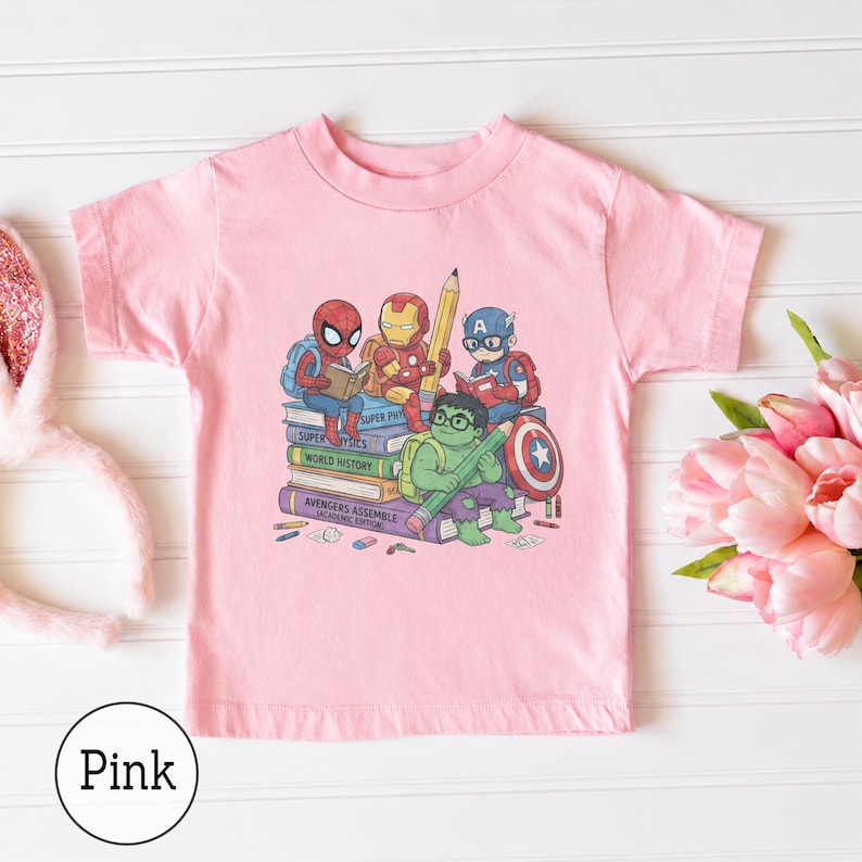 Puede incluir: Una camiseta rosa claro de manga corta con un estampado gr&aacute;fico de superh&eacute;roes de dibujos animados sentados sobre una pila de libros. Los personajes incluyen a Spider-Man, Iron Man, Captain America y Hulk, con texto en los lomos de los libros que dice Super Physics, World History y Avengers Assemble Academic Edition.