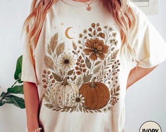 May include: An ivory short-sleeve t-shirt featuring a graphic print of two pumpkins surrounded by various wildflowers, wheat stalks, and a crescent moon. The design uses a warm color palette of brown, tan, and cream tones, centered on the chest.