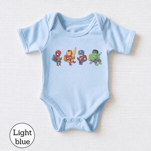 May include: A light blue short-sleeve bodysuit featuring a graphic print of four cartoon superheroes. The characters include Spider-Man reading a book, Iron Man holding a large pencil, Captain America reading, and the Hulk holding a pencil.