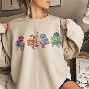 May include: A sand-colored crewneck sweatshirt featuring a graphic print of four cartoon superheroes as students. The design shows Spider-Man reading a book, Iron Man holding a large pencil, Captain America reading, and the Hulk holding a pencil.