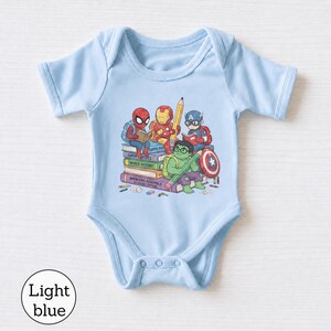 Puede incluir: Un body de manga corta color azul claro con un estampado gr&aacute;fico de Spider-Man, Iron Man, Captain America y Hulk sentados sobre una pila de libros. Los libros est&aacute;n etiquetados como Super Physics, World History y Avengers Assemble Academic Edition.