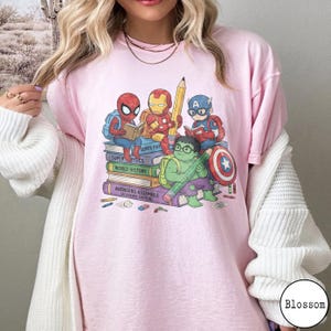 Puede incluir: Una camiseta rosa claro con un estampado gr&aacute;fico de superh&eacute;roes de dibujos animados, incluidos Spider-Man, Iron Man, Captain America y Hulk, sentados sobre una pila de libros. Los libros est&aacute;n etiquetados como Super Physics, World History y Avengers Assemble.