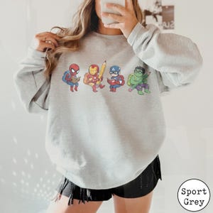 May include: A light grey crewneck sweatshirt featuring a graphic print of four cartoon superheroes. Spider-Man, Iron Man, Captain America, and the Hulk are depicted wearing backpacks and holding school supplies like books and a large yellow pencil.