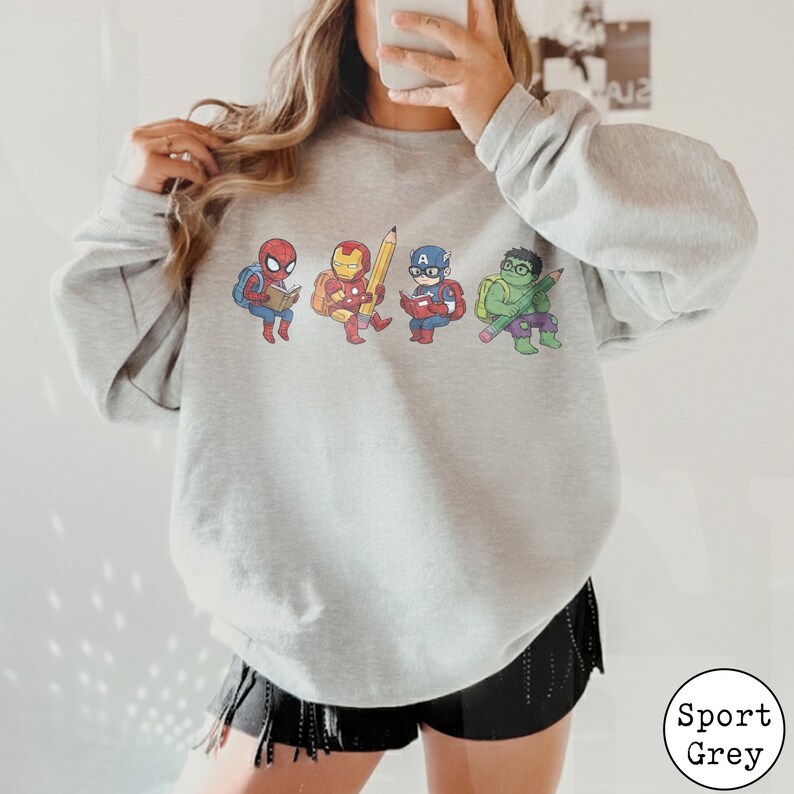 May include: A light grey crewneck sweatshirt featuring a graphic print of four cartoon superheroes. Spider-Man, Iron Man, Captain America, and the Hulk are depicted wearing backpacks and holding school supplies like books and a large yellow pencil.