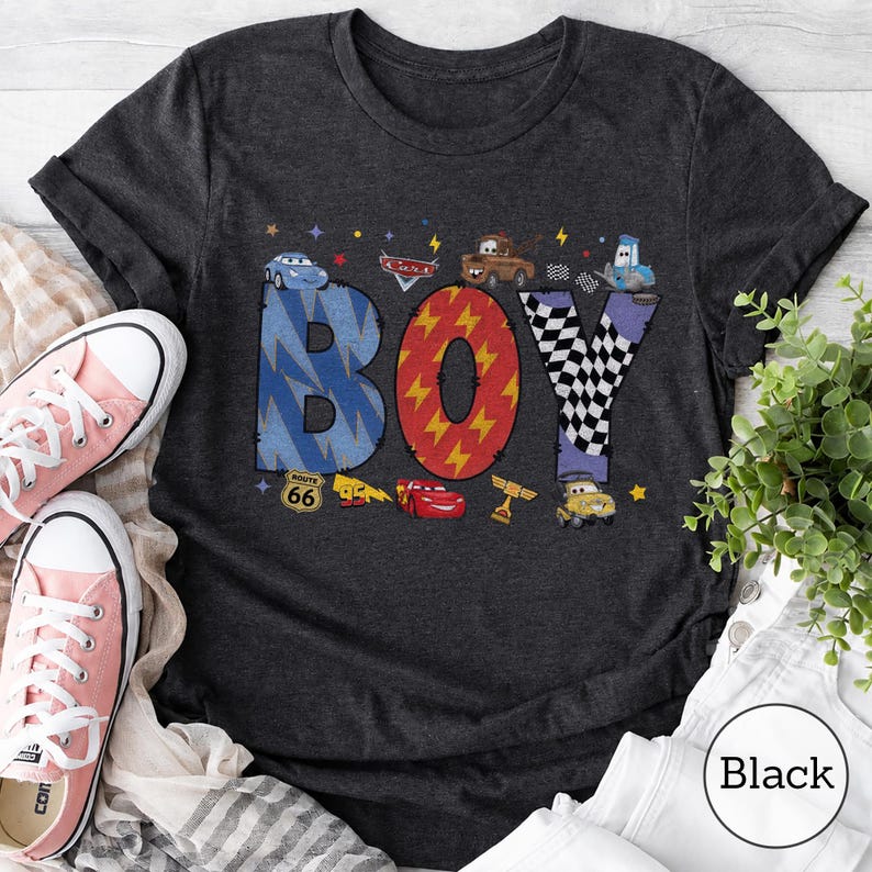 May include: A dark gray short-sleeve t-shirt featuring the word BOY in large letters. The B is blue with a lightning pattern, the O is red with yellow lightning bolts, and the Y is black and white checkered. Cars characters are printed around.