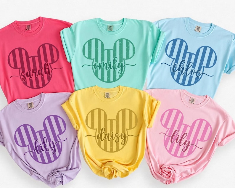 K&ouml;nnte beinhalten: Sechs kurz&auml;rmelige T-Shirts mit Rundhalsausschnitt in verschiedenen Pastellfarben wie Pink, Mintgr&uuml;n, Hellblau, Lavendel, Gelb und Hellrosa. Jedes T-Shirt zeigt eine gro&szlig;e, gestreifte Mickey-Maus-Kopf-Silhouette mit einem Namen in Schreibschrift.