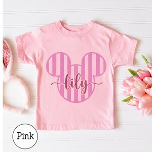 K&ouml;nnte beinhalten: Ein hellrosa T-Shirt mit kurzen &Auml;rmeln und einer mittigen Grafik einer Mausohren-Silhouette mit vertikalen Streifen in einem dunkleren Rosaton. Der Name lily ist in einer dunkelrosa Schreibschrift mittig auf der Grafik gedruckt.