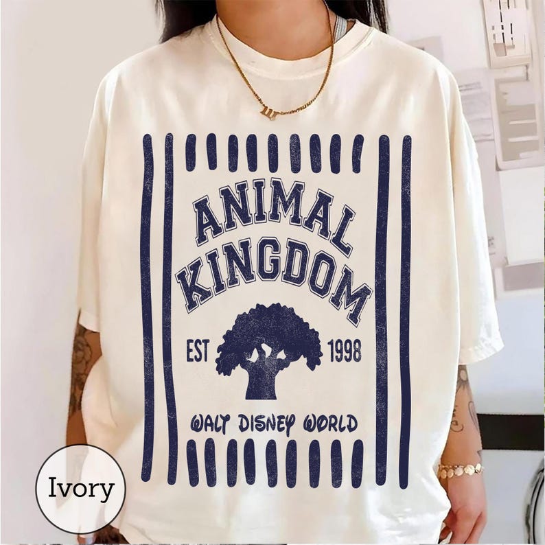 Pu&ograve; includere: Una t-shirt a maniche corte color avorio con una stampa grafica blu scuro. Il design include strisce verticali, il testo ANIMAL KINGDOM, la sagoma di un albero, EST 1998 e WALT DISNEY WORLD. La maglietta ha un girocollo e una vestibilit&agrave; ampia.