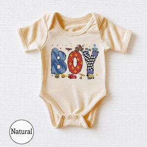 May include: A cream-colored short-sleeve bodysuit featuring a graphic print of the word BOY in large letters. The letters are decorated with blue lightning, red lightning bolts, and black and white checkered patterns, surrounded by small car characters.