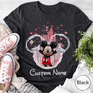 May include: A dark gray t-shirt featuring a graphic of a Mickey Mouse balloon character centered within a white outline of mouse ears. The design includes a castle silhouette, stars, and hearts in pink and red, with the text Custom Name below.