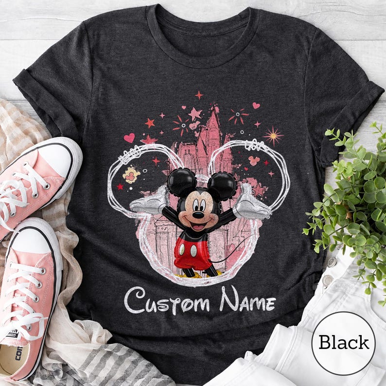 May include: A dark gray t-shirt featuring a graphic of a Mickey Mouse balloon character centered within a white outline of mouse ears. The design includes a castle silhouette, stars, and hearts in pink and red, with the text Custom Name below.
