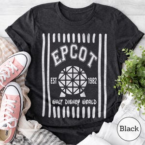Pu&ograve; includere: Una t-shirt a maniche corte grigio antracite con una stampa grafica bianca. Il design include il testo EPCOT, EST 1982 e WALT DISNEY WORLD, centrato attorno a un'icona a sfera geometrica, incorniciato da strisce verticali bianche.