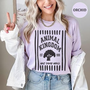 Pu&ograve; includere: Una t-shirt viola chiaro con una grafica nera con il testo ANIMAL KINGDOM, EST 1998 e WALT DISNEY WORLD, centrata sopra la sagoma di un albero, incorniciata da strisce verticali nere, indossata sotto una camicia bianca e jeans blu.