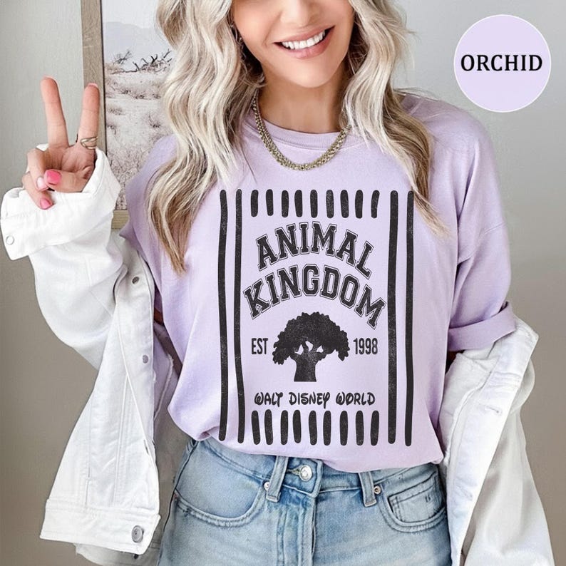 Pu&ograve; includere: Una t-shirt viola chiaro con una grafica nera con il testo ANIMAL KINGDOM, EST 1998 e WALT DISNEY WORLD, centrata sopra la sagoma di un albero, incorniciata da strisce verticali nere, indossata sotto una camicia bianca e jeans blu.