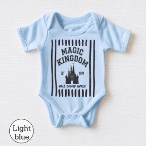 Pu&ograve; includere: Un body per neonati azzurro con una stampa grafica nera della sagoma di un castello. Il testo recita MAGIC KINGDOM, EST 1971 e WALT DISNEY WORLD, incorniciato da strisce verticali nere. Il capo presenta maniche corte e chiusura a bottoni.