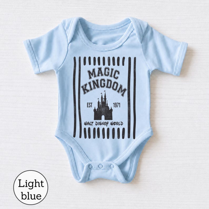Pu&ograve; includere: Un body per neonati azzurro con una stampa grafica nera della sagoma di un castello. Il testo recita MAGIC KINGDOM, EST 1971 e WALT DISNEY WORLD, incorniciato da strisce verticali nere. Il capo presenta maniche corte e chiusura a bottoni.