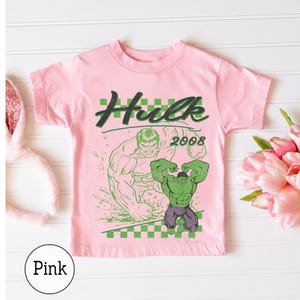 Pu&ograve; includere: Una t-shirt a maniche corte rosa chiaro con una stampa grafica verde del personaggio Hulk, il testo "Hulk" in un carattere stilizzato, l'anno "2008" e un bordo a scacchi verdi e bianchi nella parte superiore e inferiore del disegno.