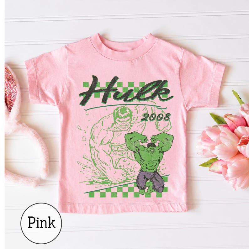 Pu&ograve; includere: Una t-shirt a maniche corte rosa chiaro con una stampa grafica verde del personaggio Hulk, il testo "Hulk" in un carattere stilizzato, l'anno "2008" e un bordo a scacchi verdi e bianchi nella parte superiore e inferiore del disegno.