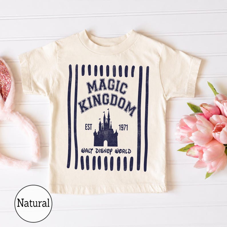 Pu&ograve; includere: Una t-shirt a maniche corte color naturale con una stampa grafica blu scuro. Il design include strisce verticali, il testo MAGIC KINGDOM, EST 1971, WALT DISNEY WORLD e la sagoma di un castello al centro della t-shirt.