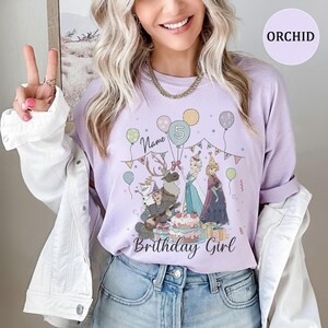Op de afbeelding: Een orchideekleurig T-shirt met een grafische print van animatiefiguren, een verjaardagstaart, ballonnen en slingers. De tekst luidt Name en Birthday Girl, met een cijfer 5 in een ballon. Het shirt heeft een ronde hals en korte mouwen.