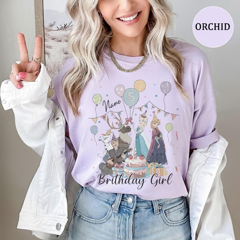 Op de afbeelding: Een orchideekleurig T-shirt met een grafische print van animatiefiguren, een verjaardagstaart, ballonnen en slingers. De tekst luidt Name en Birthday Girl, met een cijfer 5 in een ballon. Het shirt heeft een ronde hals en korte mouwen.