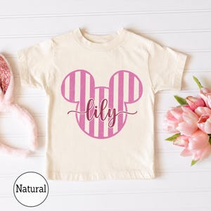K&ouml;nnte beinhalten: Ein naturfarbenes Kurzarm-T-Shirt mit einer pink-wei&szlig; gestreiften Mausohren-Silhouette als Grafik auf der Brust. Der Name lily ist in einer dunkelpinken Schreibschrift mittig &uuml;ber die Mausohren-Grafik gedruckt.