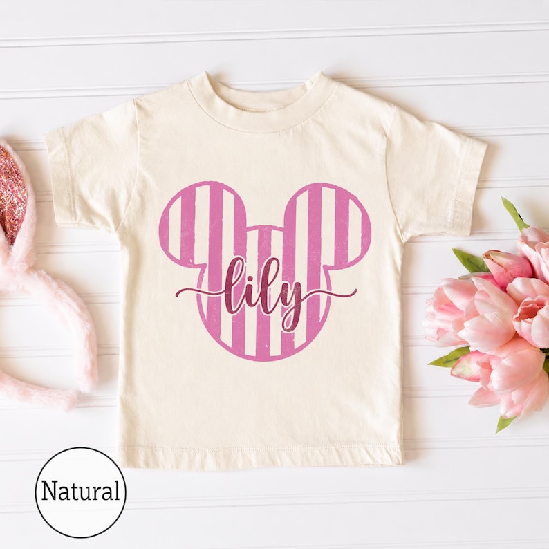 K&ouml;nnte beinhalten: Ein naturfarbenes Kurzarm-T-Shirt mit einer pink-wei&szlig; gestreiften Mausohren-Silhouette als Grafik auf der Brust. Der Name lily ist in einer dunkelpinken Schreibschrift mittig &uuml;ber die Mausohren-Grafik gedruckt.