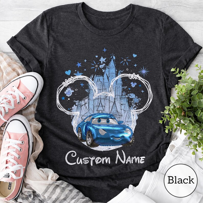 K&ouml;nnte beinhalten: Ein dunkelgraues T-Shirt mit einer wei&szlig;en Umrisszeichnung von Mausohren, die ein blaues Cartoon-Rennauto und ein Schloss umgeben. Das Design enth&auml;lt blaue Sterne und Herzen sowie den Schriftzug Custom Name in wei&szlig;er Schreibschrift.