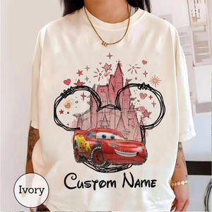 K&ouml;nnte beinhalten: Ein elfenbeinfarbenes T-Shirt mit einem Grafikdruck eines roten Rennwagens mit der Nummer 95 vor der Umrisszeichnung eines rosa Schlosses. Das Design enth&auml;lt handgezeichnete Micky-Maus-Ohren, Sterne, Herzen und den Text Custom Name in schwarzer Schrift.