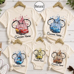 K&ouml;nnte beinhalten: Eine Kollektion aus f&uuml;nf T-Shirts und einem Body in Naturfarbe mit einer Grafik einer Schloss-Silhouette innerhalb einer Mickey-Maus-Ohren-Kontur. Jedes Kleidungsst&uuml;ck zeigt eine andere Figur aus dem Film Cars und den Text Custom Name.