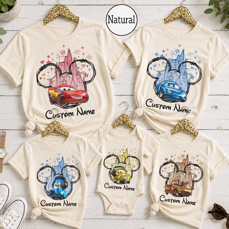 K&ouml;nnte beinhalten: Eine Kollektion aus f&uuml;nf T-Shirts und einem Body in Naturfarbe mit einer Grafik einer Schloss-Silhouette innerhalb einer Mickey-Maus-Ohren-Kontur. Jedes Kleidungsst&uuml;ck zeigt eine andere Figur aus dem Film Cars und den Text Custom Name.