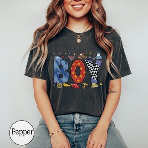 May include: A charcoal gray t-shirt featuring the word BOY in large, patterned letters. The B is blue with lightning bolts, the O is red with yellow lightning, and the Y is black and white checkered. Includes small car character graphics.
